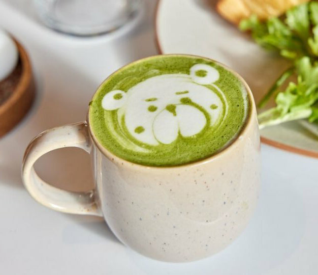 A Bondi Beach Tea Co Organic Japanese Matcha Latte, with a cute bear face latte art, rests on a table. A plate of greens and bread is visible nearby, highlighting both the artistic appeal and health benefits.