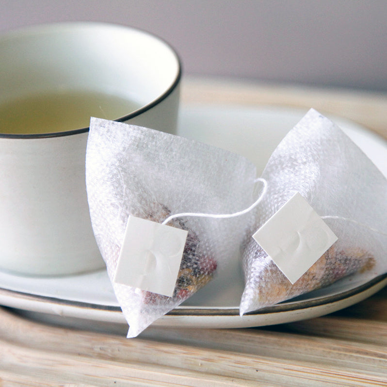 A cup of tea sits on a white saucer beside two pyramid-shaped Essiac Tea Blend - Organic Tea Bags by Bondi Beach Tea Co, on a wooden surface. The light color hints at a wellness blend focused on detoxification.