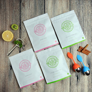Four packages of The Bondi B-Slim Together Tea by Bondi Beach Tea Co. are placed on a wooden surface, accompanied by a kiwi slice, half a lemon, mint leaves, and three cinnamon sticks. Two colorful tea infusers nearby make the perfect companions for your antioxidant-packed red tea experience.