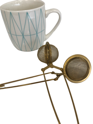 A white mug with a blue geometric design is placed next to a Bondi Beach Tea Co Clamp Style Tea Infuser, featuring dual mesh compartments, on a clean white background.