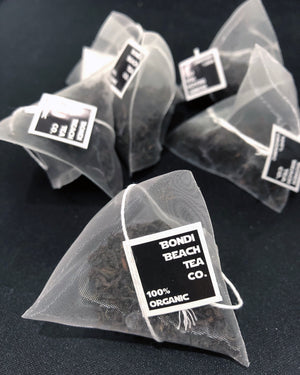 Pyramid-shaped tea bags labeled Bondi Beach Tea Co. English Breakfast Tea - Premium Organic, featuring a rich blend of Assam and Ceylon Black Tea, are scattered on a sleek black surface.