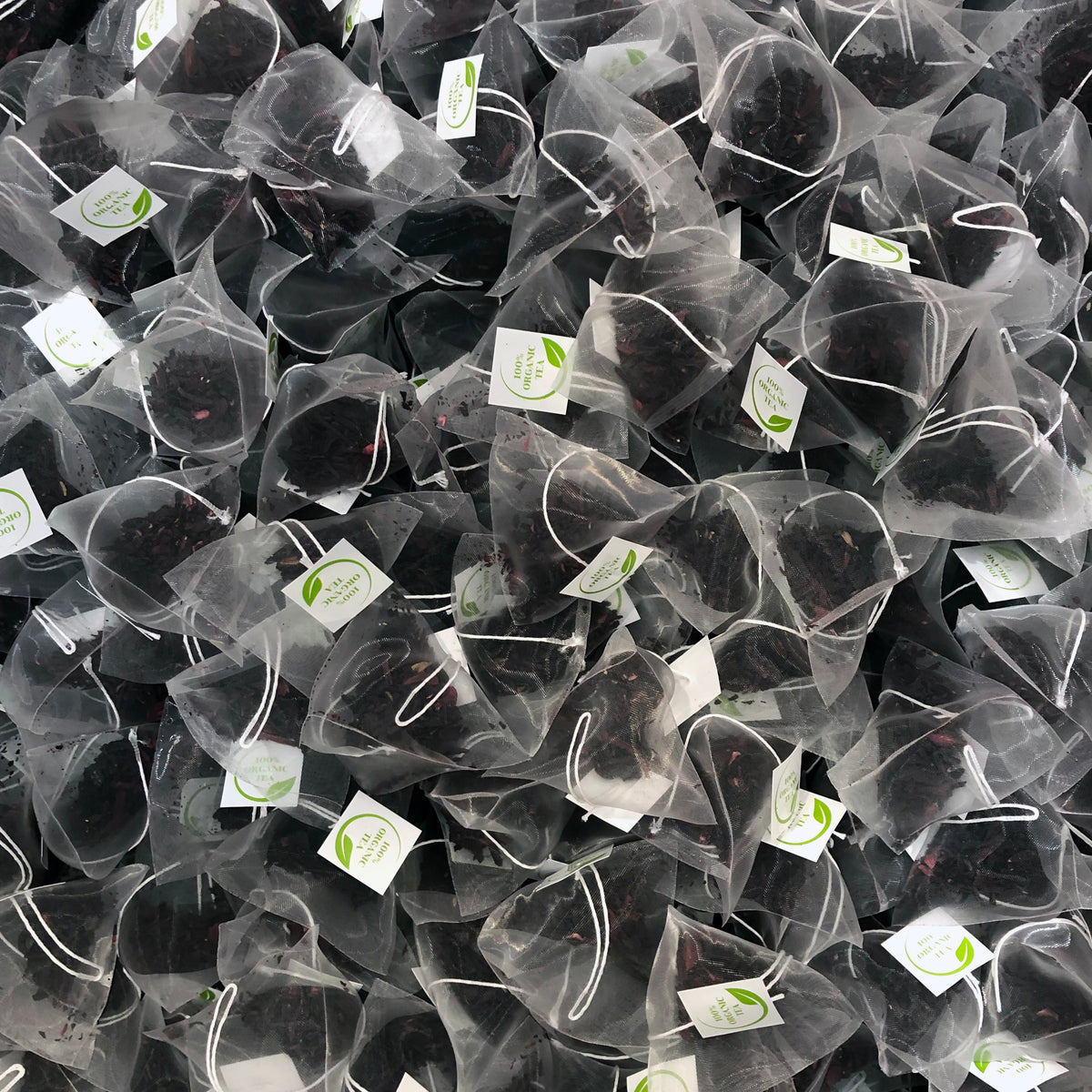 A pile of pyramid-shaped tea bags contains Premium Organic English Breakfast Tea by Bondi Beach Tea Co. Each transparent bag, with a small white tag and green logo attached by a string, reveals the rich texture and color of the tea leaves inside.