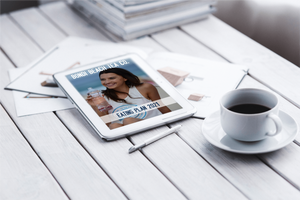 A tablet on the table displays the FREE Bondi Beach Tea Co. Eating Plan. Nearby, a woman holds a water bottle while papers, an open notebook, a pen, and a steaming white cup of coffee are scattered around.