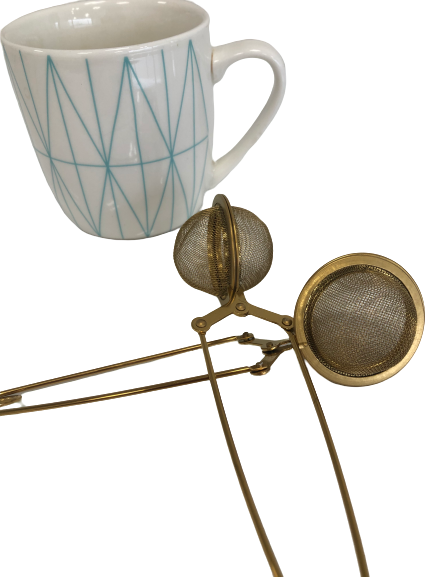 A white mug with a blue geometric design is placed next to a Bondi Beach Tea Co Clamp Style Tea Infuser, featuring dual mesh compartments, on a clean white background.