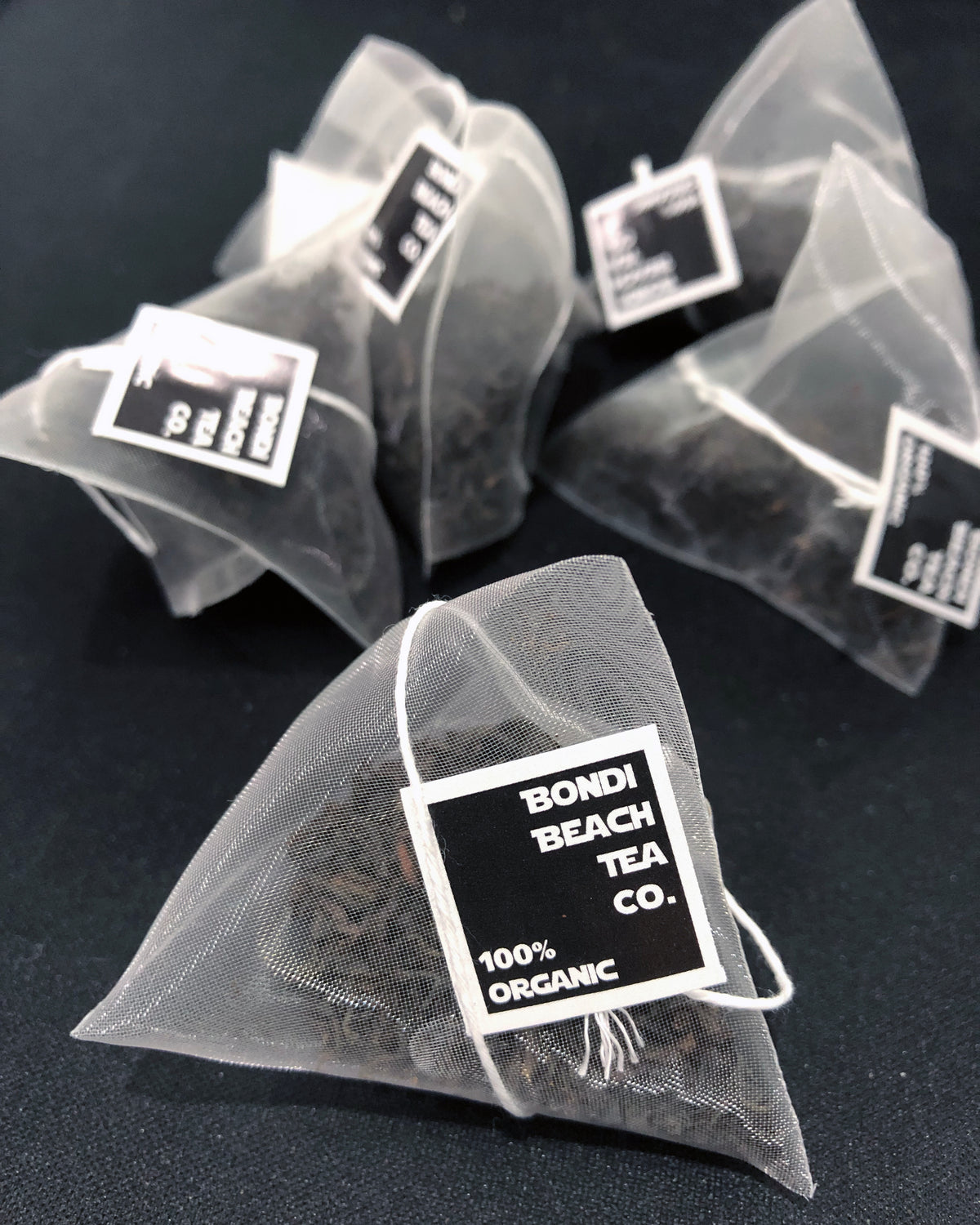 Pyramid-shaped mesh tea bags labeled Bondi Beach Tea Cos 100% Organic are scattered on a dark surface, each holding loose leaf green tea with jasmine flowers, capturing the elegance of Bondi Jasmine Tea.