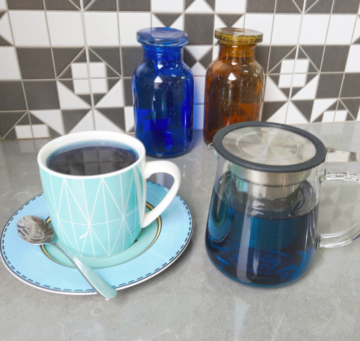 A vibrant turquoise cup of Organic Blue Butterfly Pea Flower Tea from Bondi Beach Tea Co, rich in antioxidants, rests on a matching saucer with a spoon beside a glass teapot. In the background, three colored glass jars—blue and amber—contrast against a geometric tiled wall.